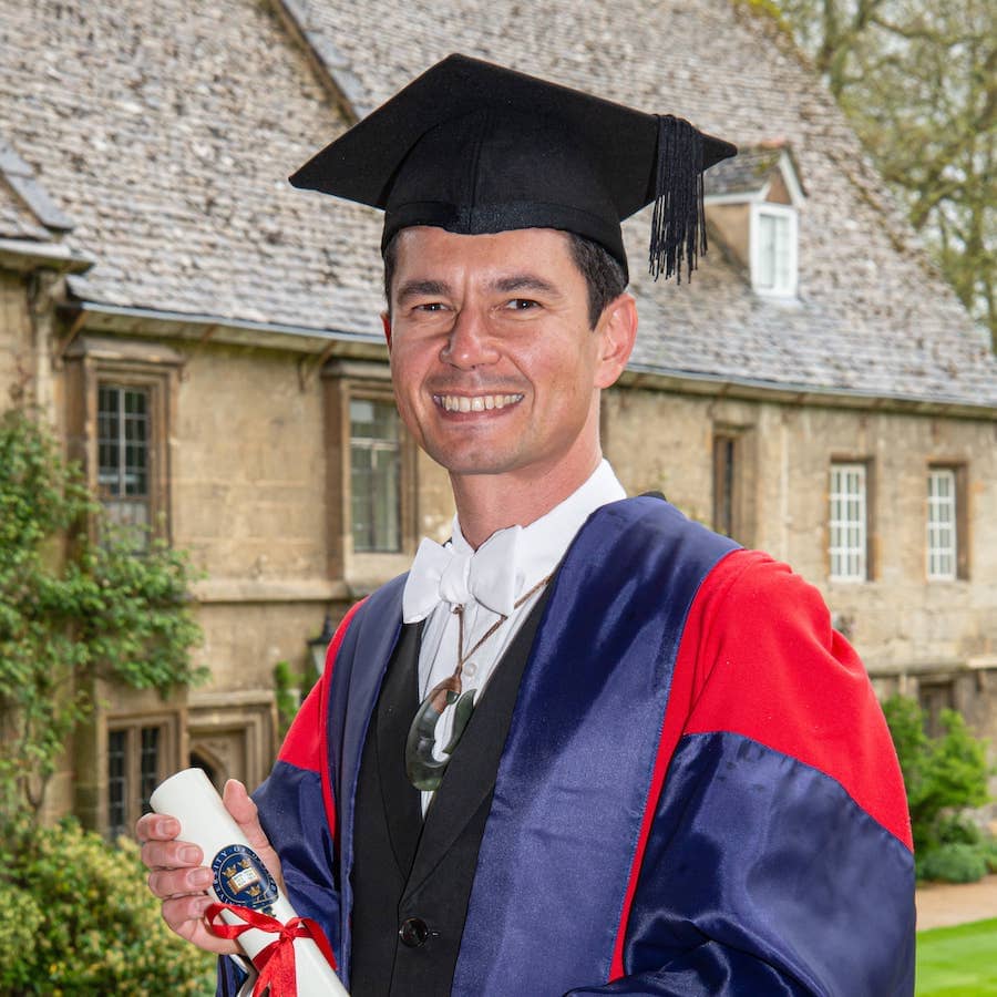 Photography of Lyndon Drake wearing his academic dress and holding his certificate of graduation.